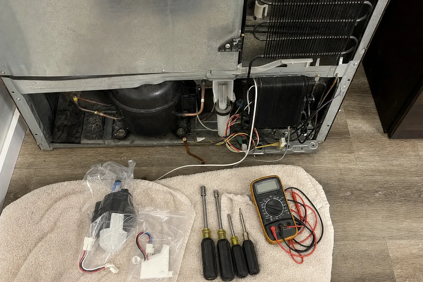 Lower grille removed from a built-in refrigerator, compressor and condenser coil exposed, replacement parts and nut drivers laid out on a towel