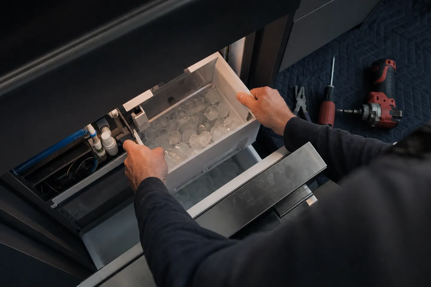 Overhead view of a clear ice bin full of round cubes lifted out of an ice-maker drawer, the blue supply line and its push-fit fittings visible in the bay