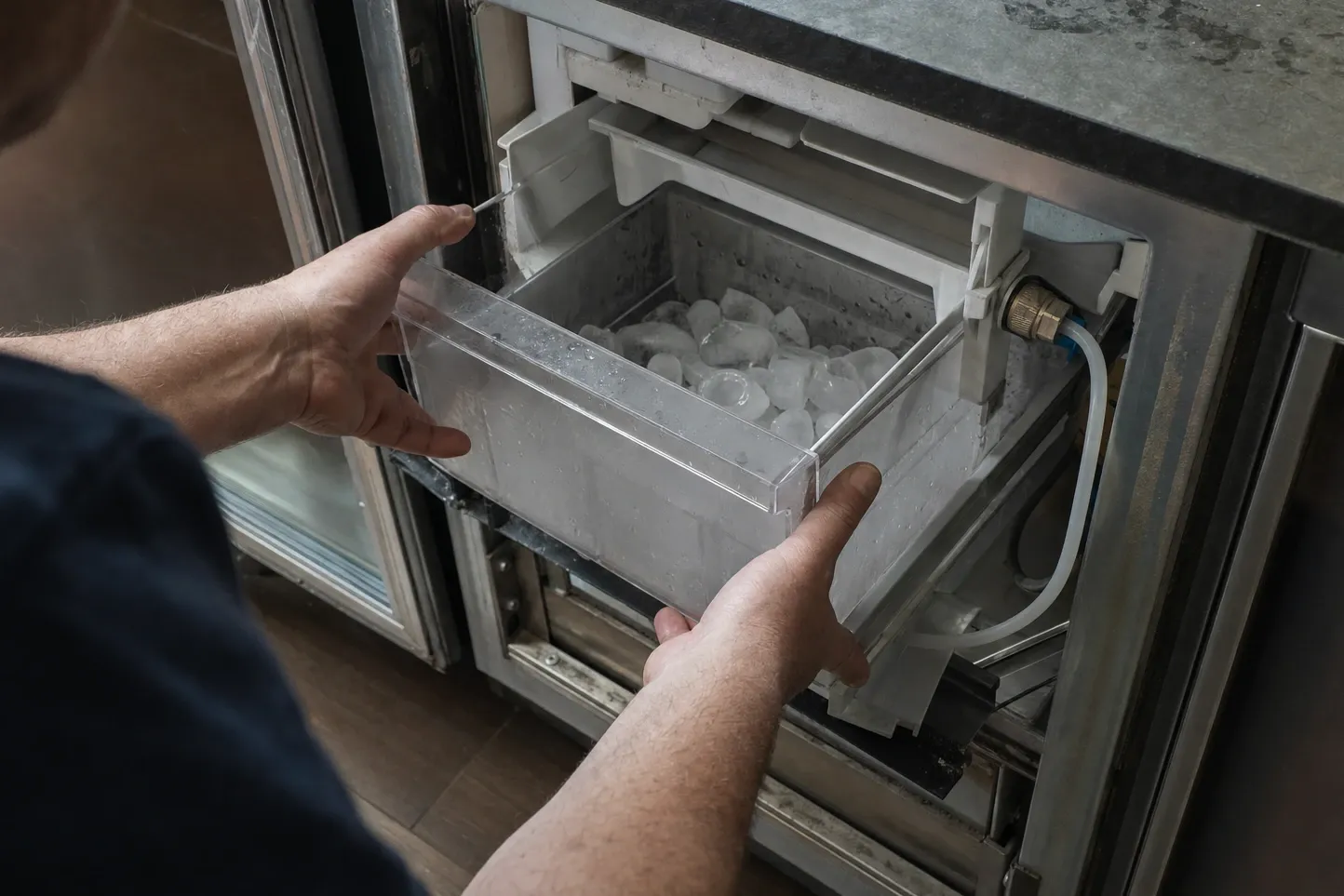 Clear ice bin drawn out of an undercounter ice maker below a stone counter, the ice mould and a brass water fitting visible in the bay behind it